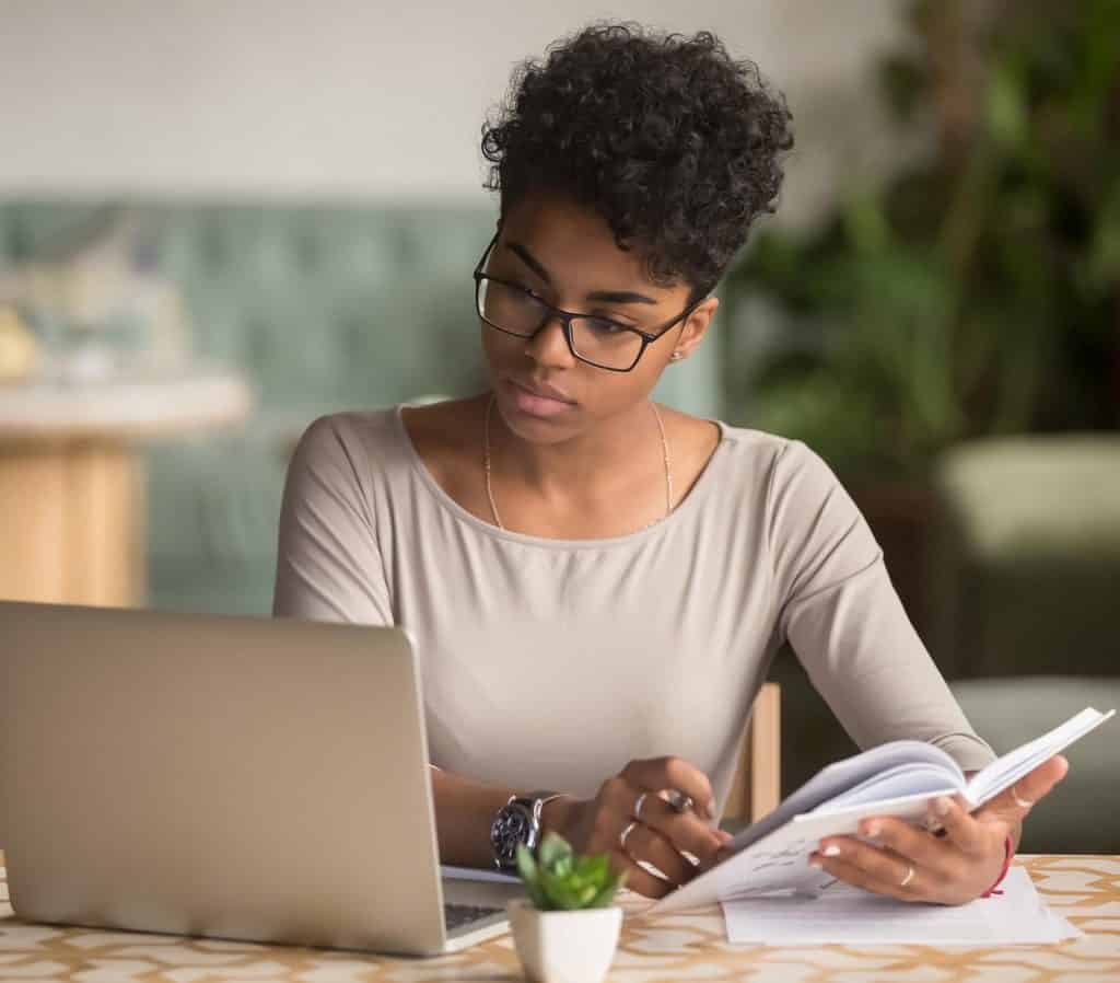 A young professional woman with glasses studies intently, cross-referencing a book with her laptop for an eLearning project.