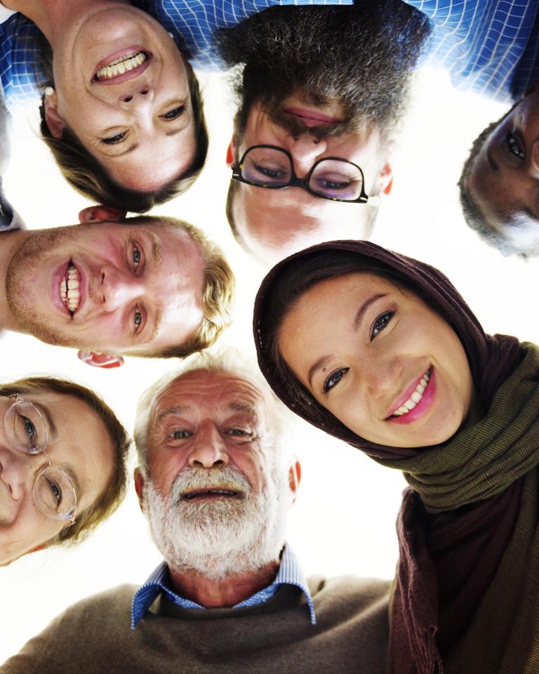 A diverse group of smiling people of different ages and ethnicities huddle together, looking down at the camera.