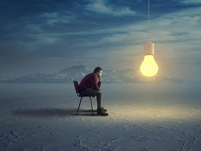A man sits in a vast landscape, contemplating a single glowing lightbulb, symbolizing a creative idea or business innovation.