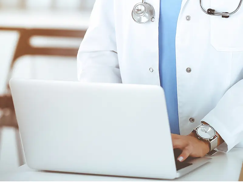 A doctor in a white lab coat with a stethoscope around their neck is typing on a laptop in a brightly lit clinic office.